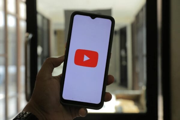 youtube news - Close-up of a hand holding a smartphone displaying the YouTube logo in a well-lit room.