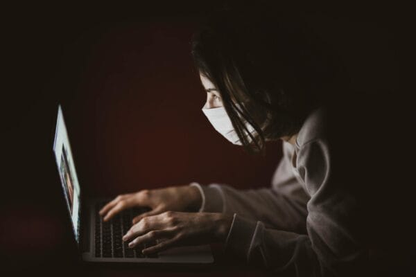 ai news today - A woman wearing a face mask focuses on her laptop screen in dim lighting, highlighting remote work.