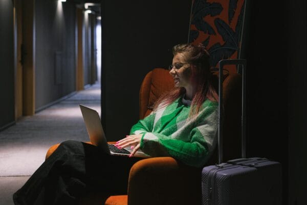 wordpress hotel theme - A woman in a hotel hallway using a laptop, sitting on an armchair next to luggage, creating a cozy working environment.