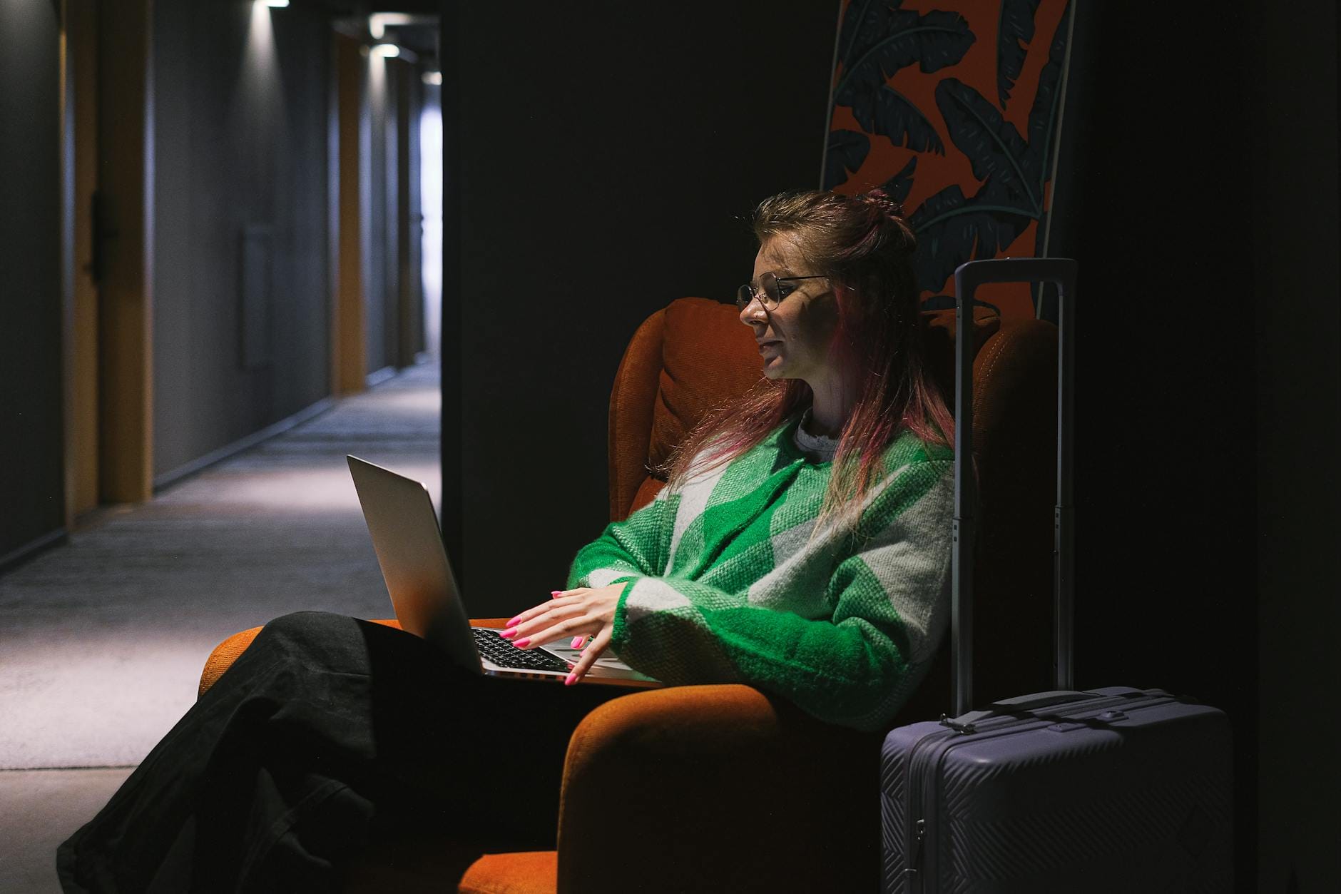 wordpress hotel theme - A woman in a hotel hallway using a laptop, sitting on an armchair next to luggage, creating a cozy working environment.
