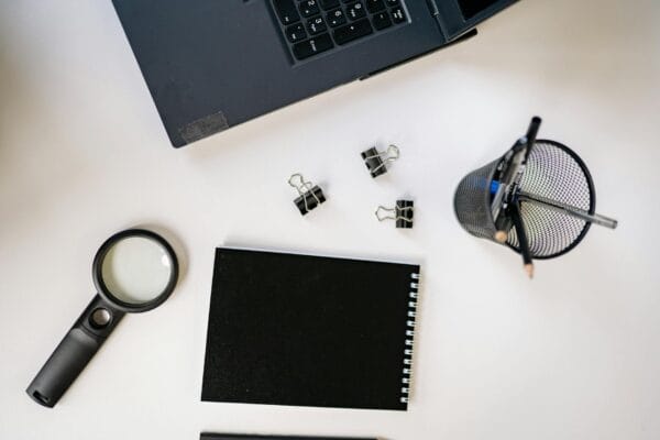 keyword research advanced - Flat lay of a workspace featuring a laptop, black notebook, magnifying glass, and office supplies on a white surface.
