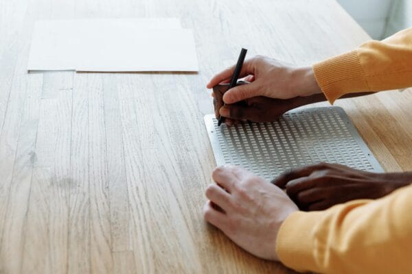AI writing assistant - Adult guiding another in learning Braille writing indoors, hands close-up.