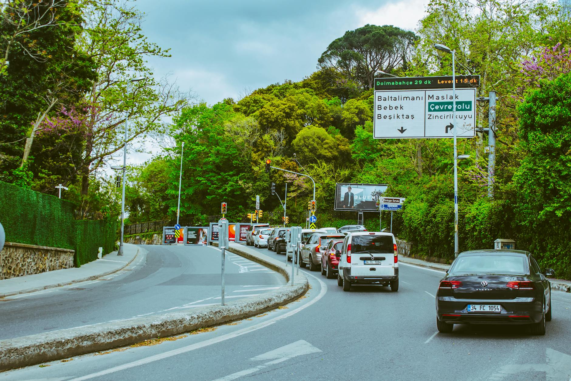 traffic organik - Cars line up on a lush green road under a cloudy sky, with prominent road signs.