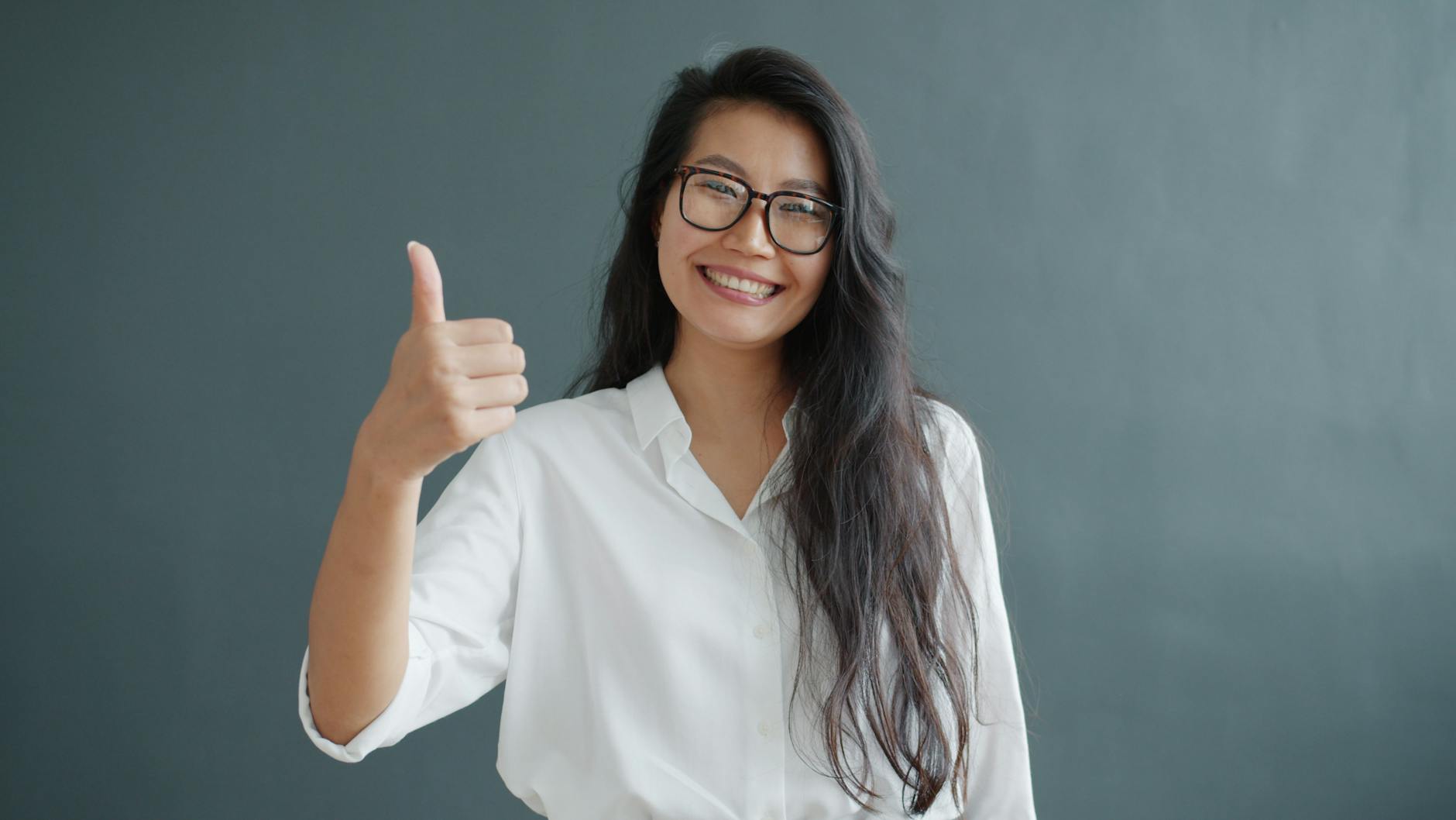 audit seo professional and optimization - Smiling woman in glasses and white shirt giving a thumbs up against gray background.
